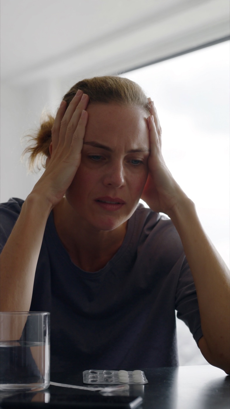 A woman with auburn hair pressing her temples during a migraine attack, a blister of pills on the table